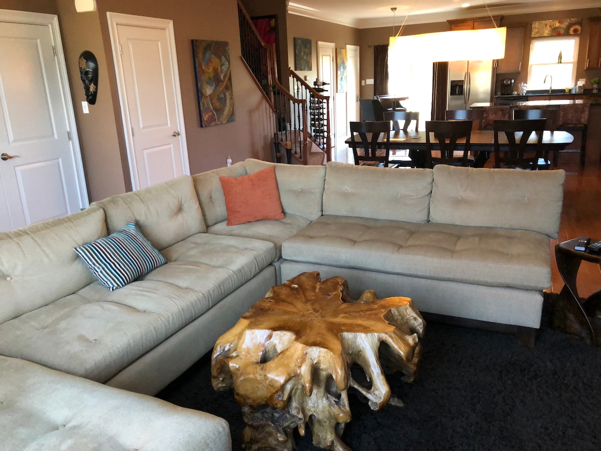 A spacious living area showcases a light-colored sectional sofa with an accent pillow, arranged around a natural wood coffee table. Warm lighting highlights the textured walls, while dining chairs and an open kitchen are visible in the background.
