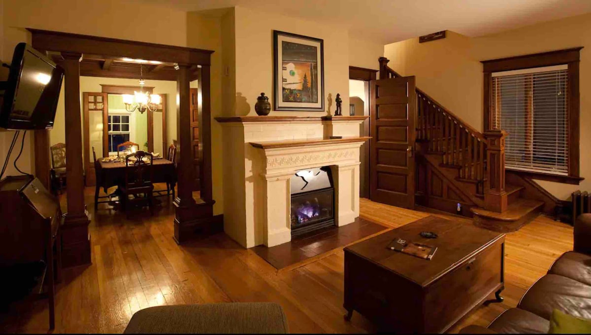 A warm living area features hardwood floors and a central fireplace. A wooden table can be seen in the adjacent dining space, while intricate wooden details and a staircase enhance the home's historic charm. Soft lighting contributes to the inviting ambiance.