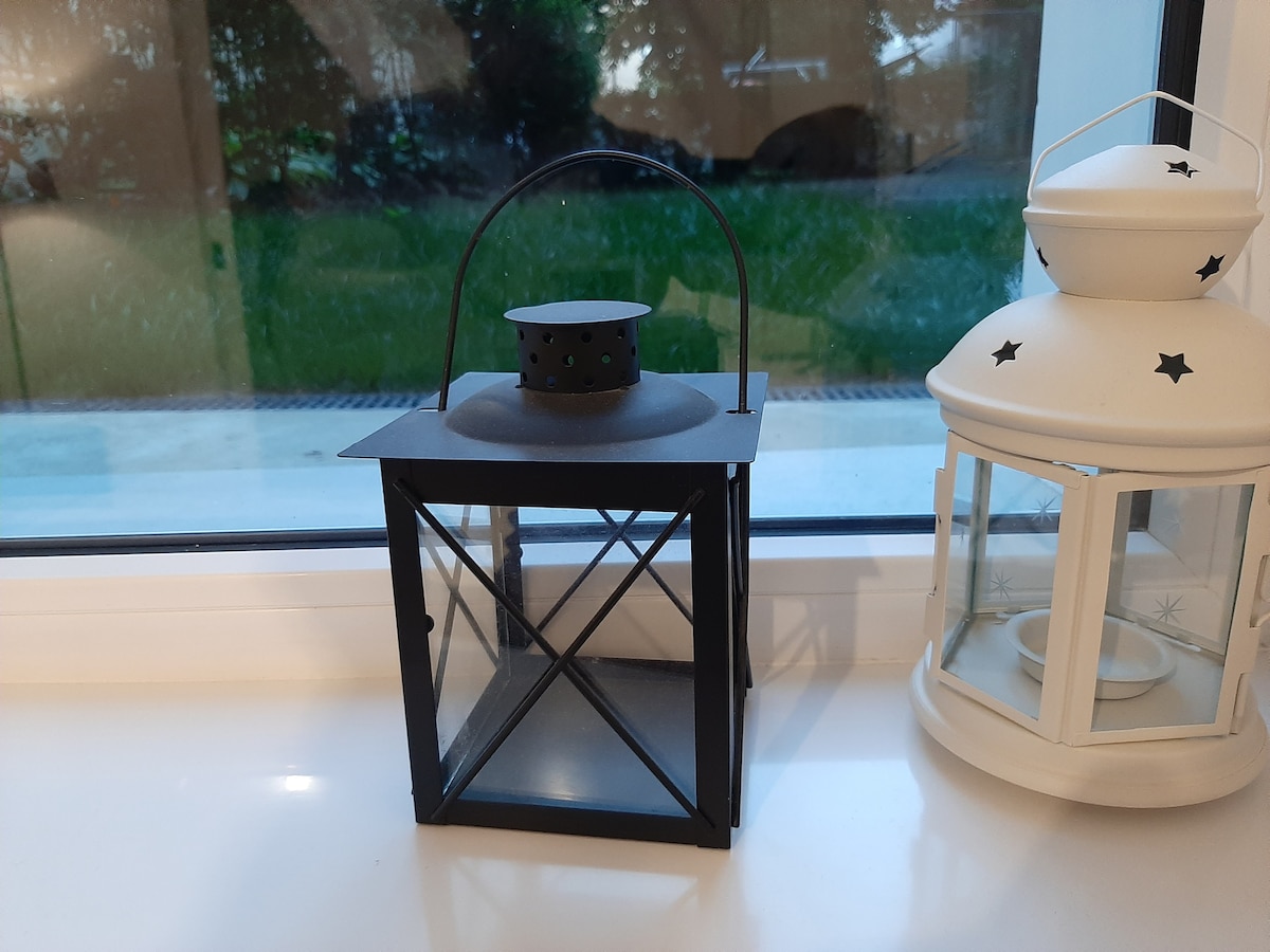 Two decorative lanterns are positioned on a windowsill. One lantern is black with a geometric design, and the other is white with star patterns. Natural light filters in from the window, illuminating the serene outdoor view of greenery beyond.