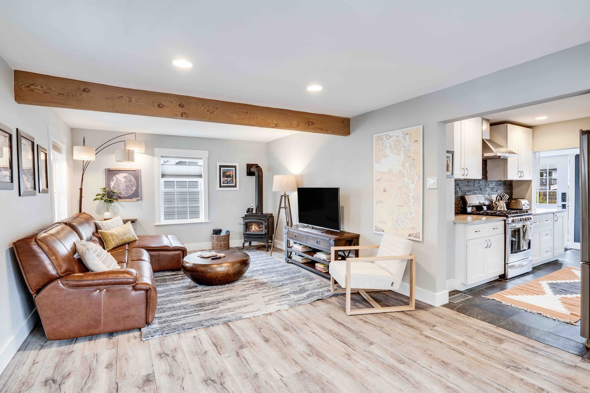 The open living area features a spacious leather sectional sofa and a round wooden coffee table on a textured rug. A television is positioned across from the seating, while a well-equipped kitchen with light cabinetry is visible in the background, complemented by a stylish light fixture.