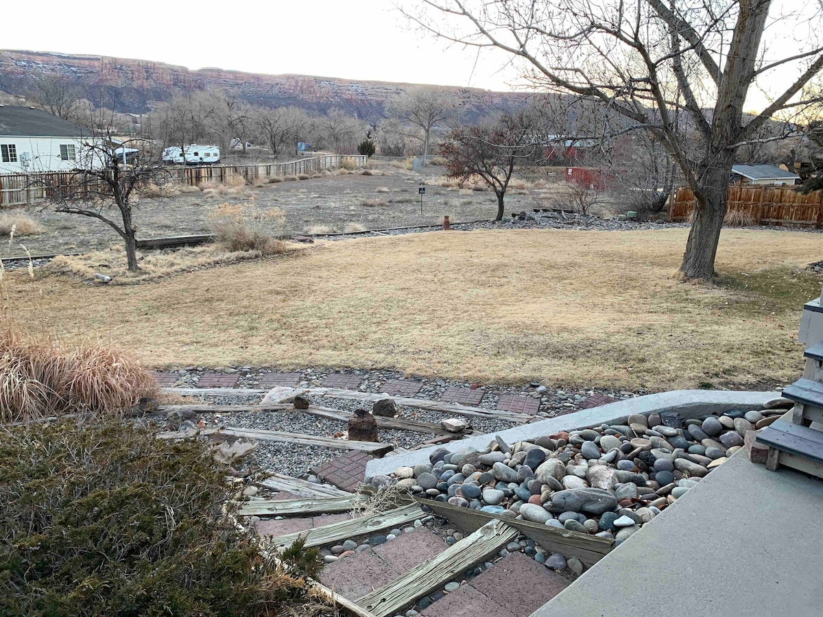 The image depicts a spacious outdoor area featuring a gently sloping lawn with natural grass. Boulders and stones line the walkways, leading to the grassy expanse. Leafless trees are present, set against a backdrop of distant hills. The sky appears clear, indicating an open atmosphere.