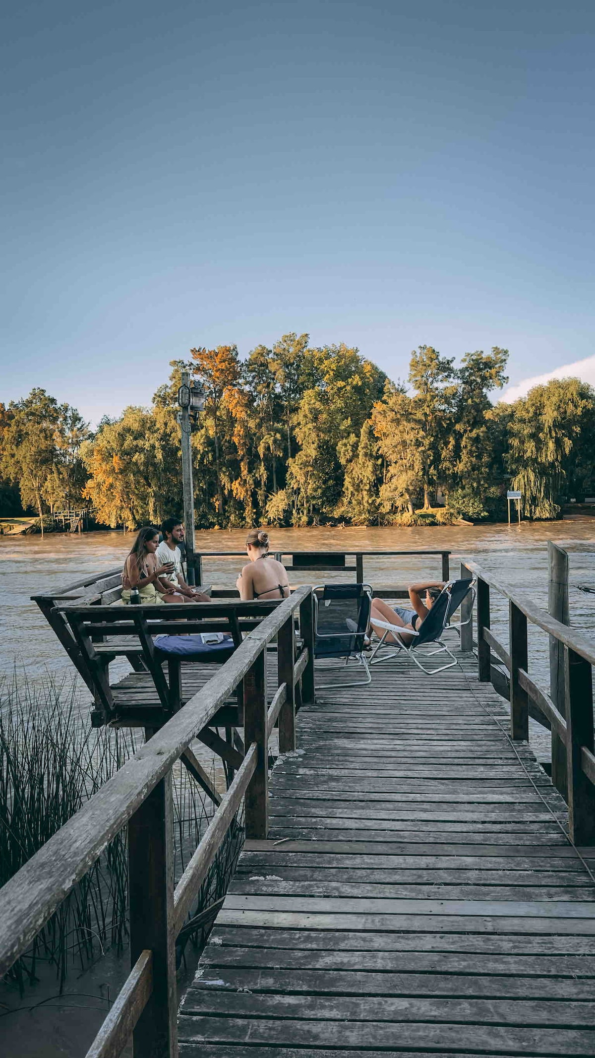 A wooden dock stretches over the water, surrounded by lush trees displaying autumn colors. Four chairs are positioned along the dock, inviting relaxation while overlooking the scenic view. The tranquil atmosphere is enhanced by the gentle ripples of the water.