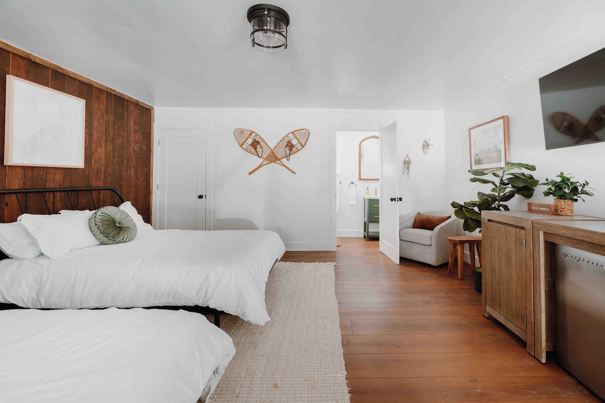 The room features two queen beds with white linens against a backdrop of wood paneling. A seating area with a small sofa is visible. Decor is complemented by wall art and a flat-screen TV. A doorway leads to a private bathroom, enhancing functional living space.