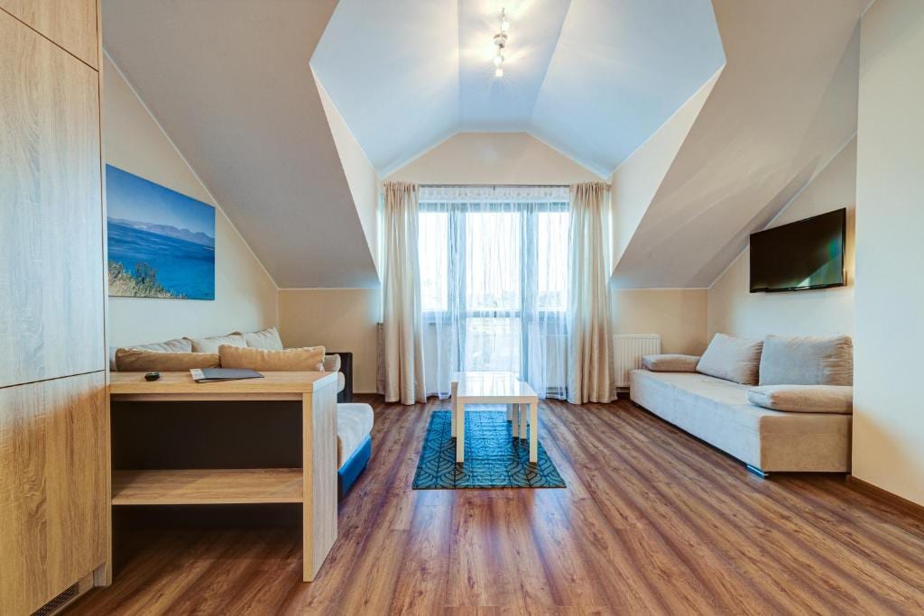 A spacious living area features sloped ceilings and large windows allowing natural light to fill the room. Neutral-toned sofas are arranged around a central coffee table. A television is mounted on the wall, and light wooden furnishings complement the warm wood flooring.