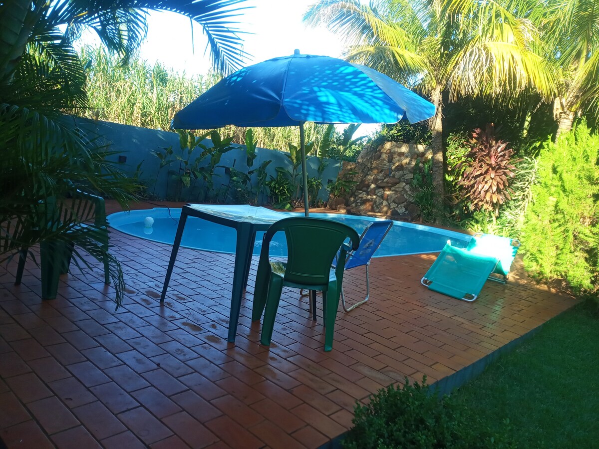 A pool area is surrounded by tropical plants, featuring a blue umbrella shading a small table with chairs. A lounge chair lies near the pool's edge, and the warm tiled patio offers a relaxed atmosphere for outdoor enjoyment.
