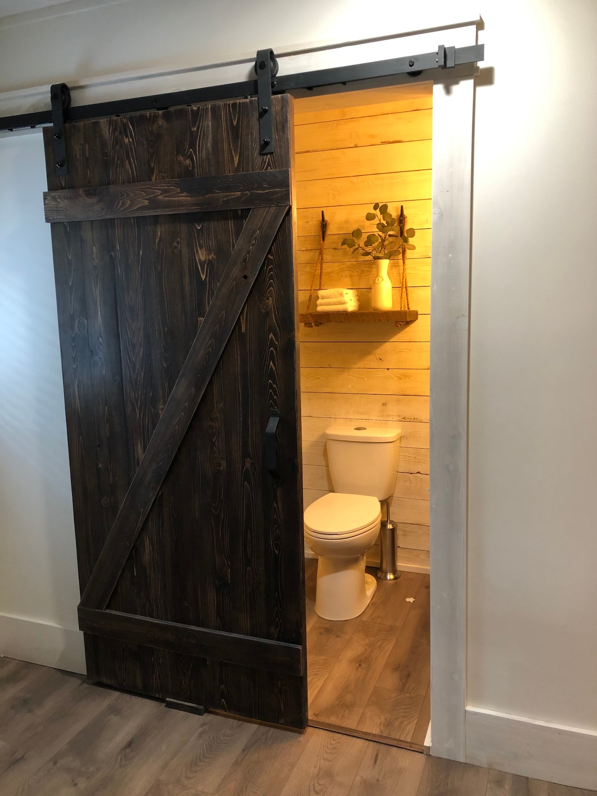 A sliding barn door reveals a bathroom space featuring wooden wall accents and warm lighting. A modern toilet is positioned adjacent to a small shelf holding decorative items. The flooring is a light wood, maintaining a cohesive style with the suite.