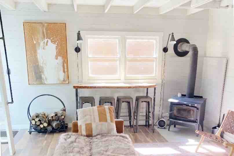 The main living area is illuminated by natural light, featuring a rustic wooden bar with stools. A wood-burning stove stands beside an arrangement of firewood. A textured artwork adorns the wall, adding visual interest to the space.