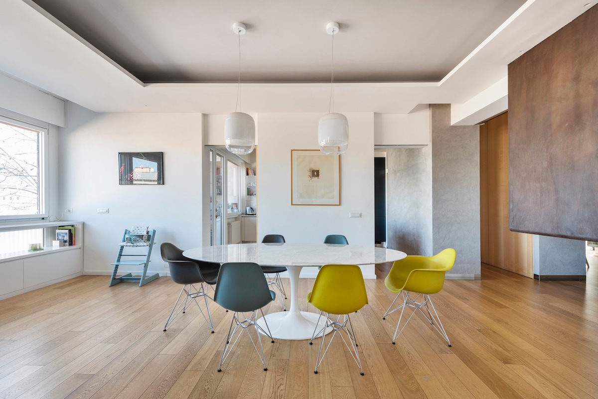 A modern dining area features a circular white table surrounded by six chairs in varying colors. Pendant lights hang above, illuminating the wooden floor. Large windows provide natural light, enhancing the spacious feel of the room.
