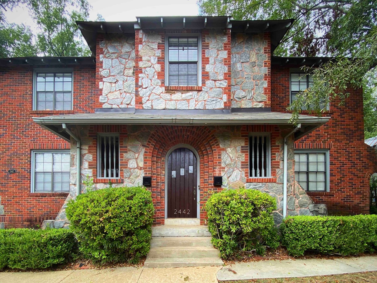 The exterior of a two-story historic building is showcased, featuring a combination of stone and brick materials. The entrance is highlighted by a large wooden door with decorative ironwork. Neatly trimmed hedges line the walkway, enhancing the inviting appearance of the residence.