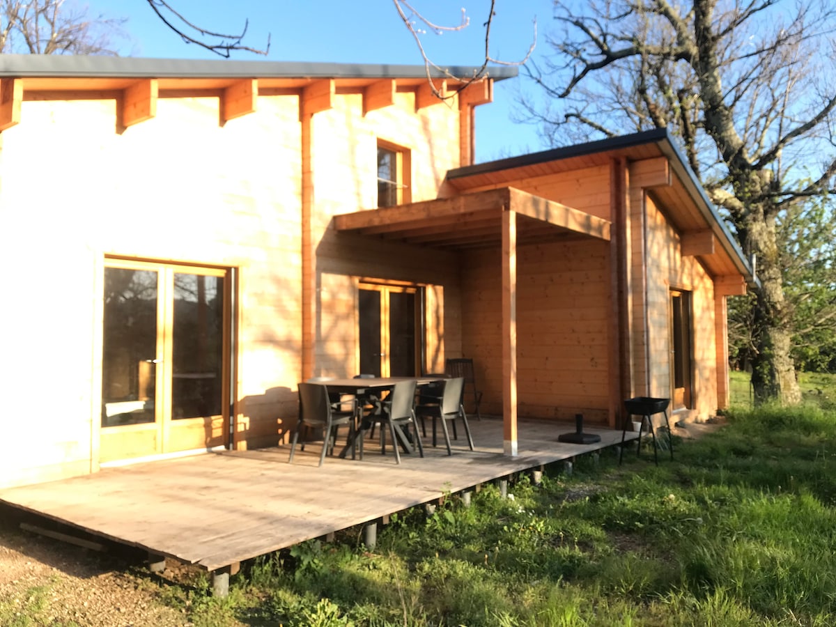A wooden chalet is seen from an outdoor patio, featuring a shaded area with a dining table and chairs set against a backdrop of trees. Large windows allow natural light to fill the interior, enhancing the cozy ambiance.