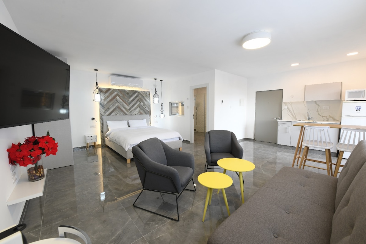 A modern suite is depicted with an open layout, featuring a spacious bed against a textured headboard. A television is mounted above a simple console. Seating includes two dark chairs around yellow tables and a light grey sofa, accompanied by a small kitchenette in the background.