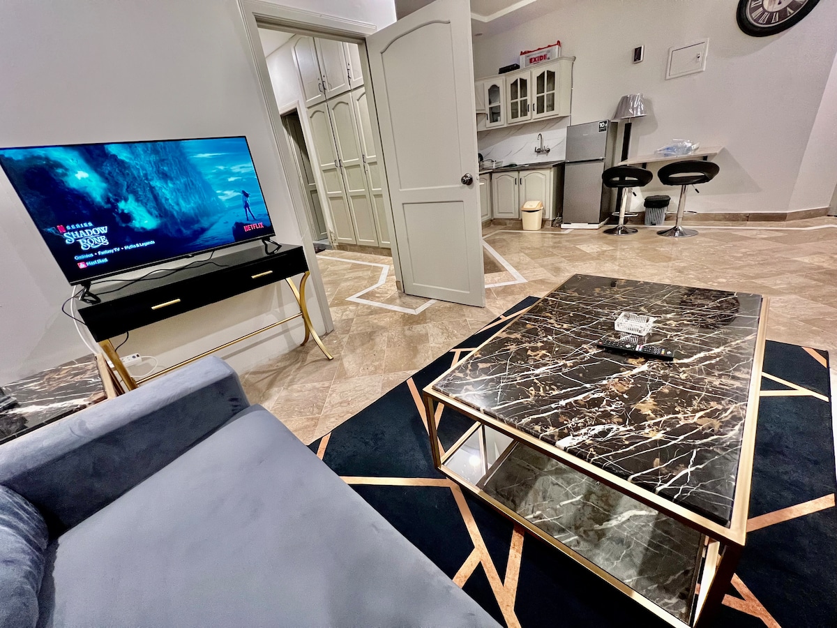 A modern living area features a sleek gray sofa positioned near a stylish coffee table with a marble surface. A television is mounted on the wall, displaying content. The space connects to a minimalist kitchen, characterized by light cabinetry and bar stools.