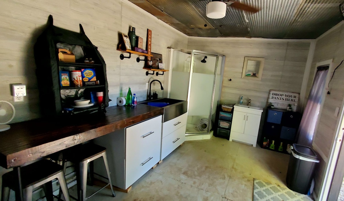 A compact kitchenette is visible, featuring a wooden countertop with bar stools. A shower area is situated in the corner, enclosed by glass. Storage cabinets and a variety of cleaning supplies are organized along the wall, contributing to a functional and efficient layout.