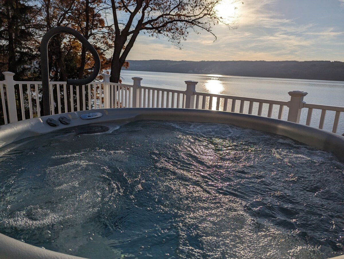 A hot tub is situated on a deck overlooking the serene waters of Seneca Lake. The water is gently disturbed, reflecting the early evening sunlight. Surrounding trees frame the scene, enhancing the tranquil ambiance.