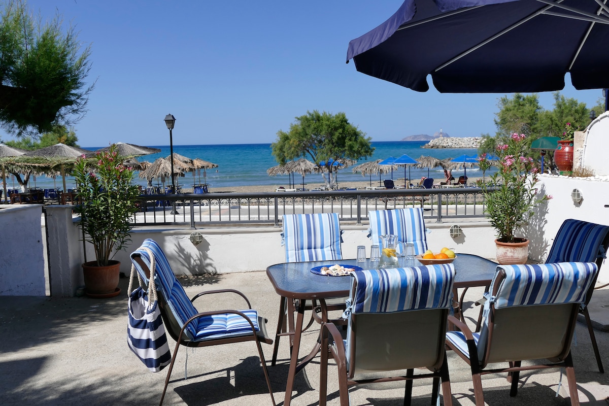 A patio area is featured with a glass-top table surrounded by striped chairs, offering a view of the sandy beach and the Libyan Sea. Sun loungers and umbrellas are visible in the background, enhancing the inviting outdoor space.