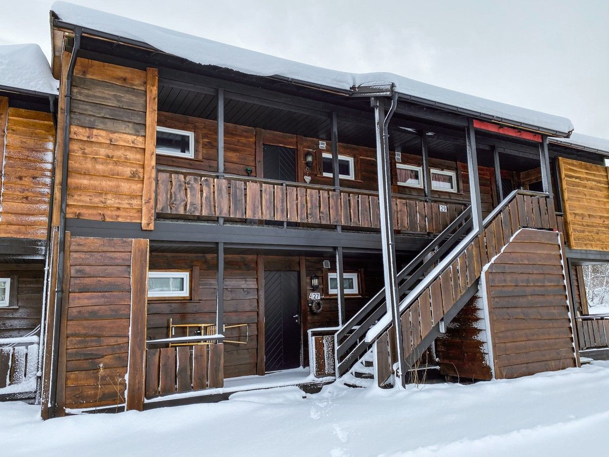 The exterior of a two-story wooden building is displayed, featuring a snow-covered entrance. A staircase leads to the upper level, while wooden railings and window frames add to the structure's rustic charm.