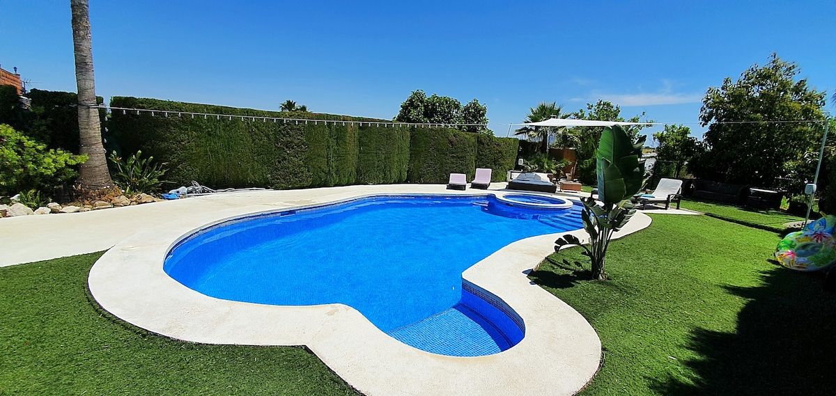 A heart-shaped swimming pool is surrounded by lush greenery and well-maintained grass. Sun loungers are positioned near the pool's edge, offering a relaxing space. A small Jacuzzi is integrated into the pool area, emphasizing the outdoor leisure environment.