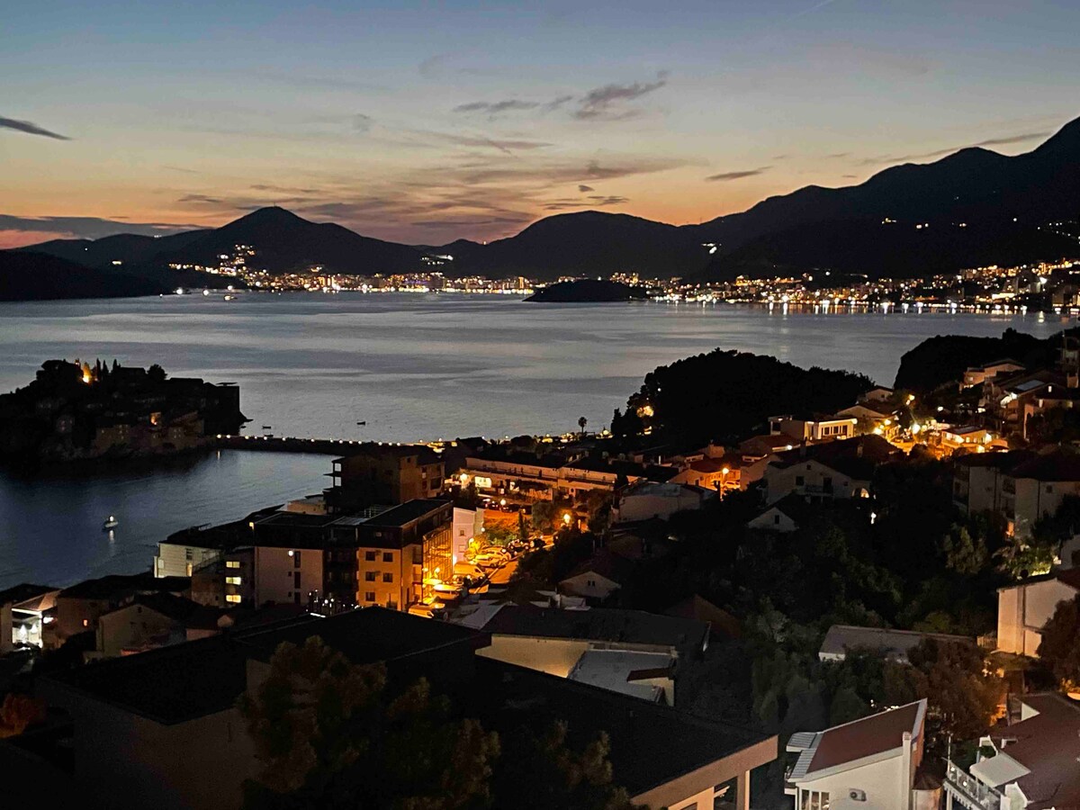 A serene view of a coastal town at dusk, with twinkling lights reflecting off the water. Mountains rise in the background, creating a peaceful backdrop for the colorful houses and shoreline. The calm water gently laps at the edge of the town, enhancing the tranquil setting.