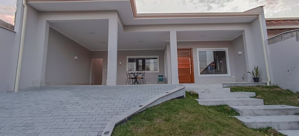 A modern home exterior features a spacious patio area with a small dining table set for four. Soft green grass borders the stone pathway leading to the entrance, which showcases a warm wooden door. Large windows allow natural light to fill the interior.