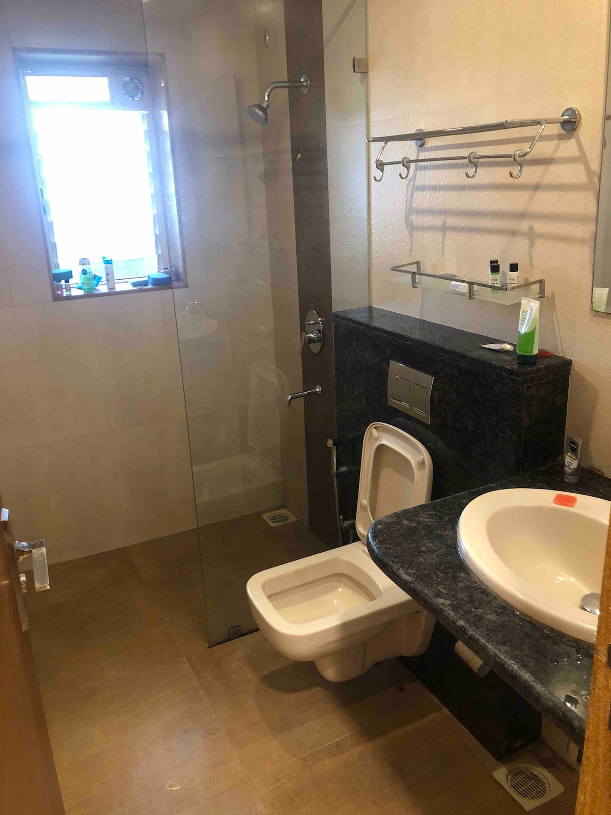 A well-equipped bathroom features a clear glass shower enclosure and a modern toilet. A round sink is set into a dark countertop, complemented by a wall-mounted rack with toiletries. Natural light enters from a small window, illuminating the neutral-toned tiles.