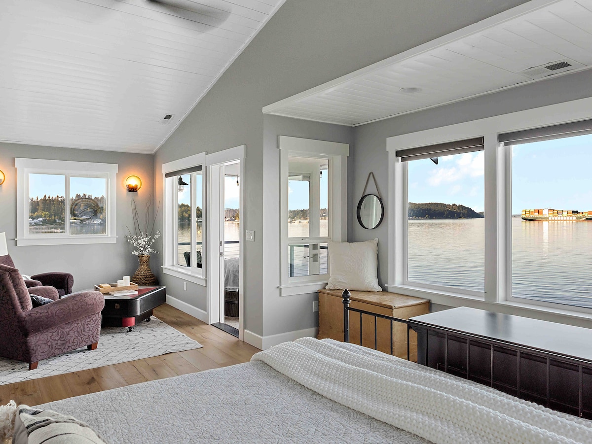 A serene master suite features large windows offering stunning water views. The space includes a cozy sitting area with a plush armchair and a coffee table, a neatly made bed, and warm wooden accents. Natural light brightens the room, enhancing the tranquil atmosphere.