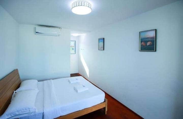 A bright and airy sleeping area features a spacious bed with white linens neatly arranged. The light-colored walls complement the warm wooden flooring. Two framed artworks are displayed on the wall, and a window allows natural light to fill the room.