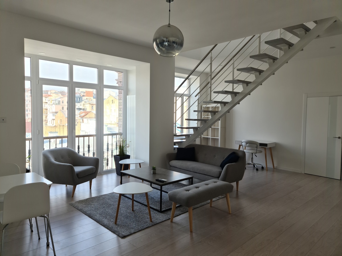 A bright, spacious living area is illuminated by large windows, offering views of the surrounding city. A modern staircase leads to an upper level. Comfortable seating options include a sofa and armchairs, complemented by a coffee table and a rug on the light wooden floor.