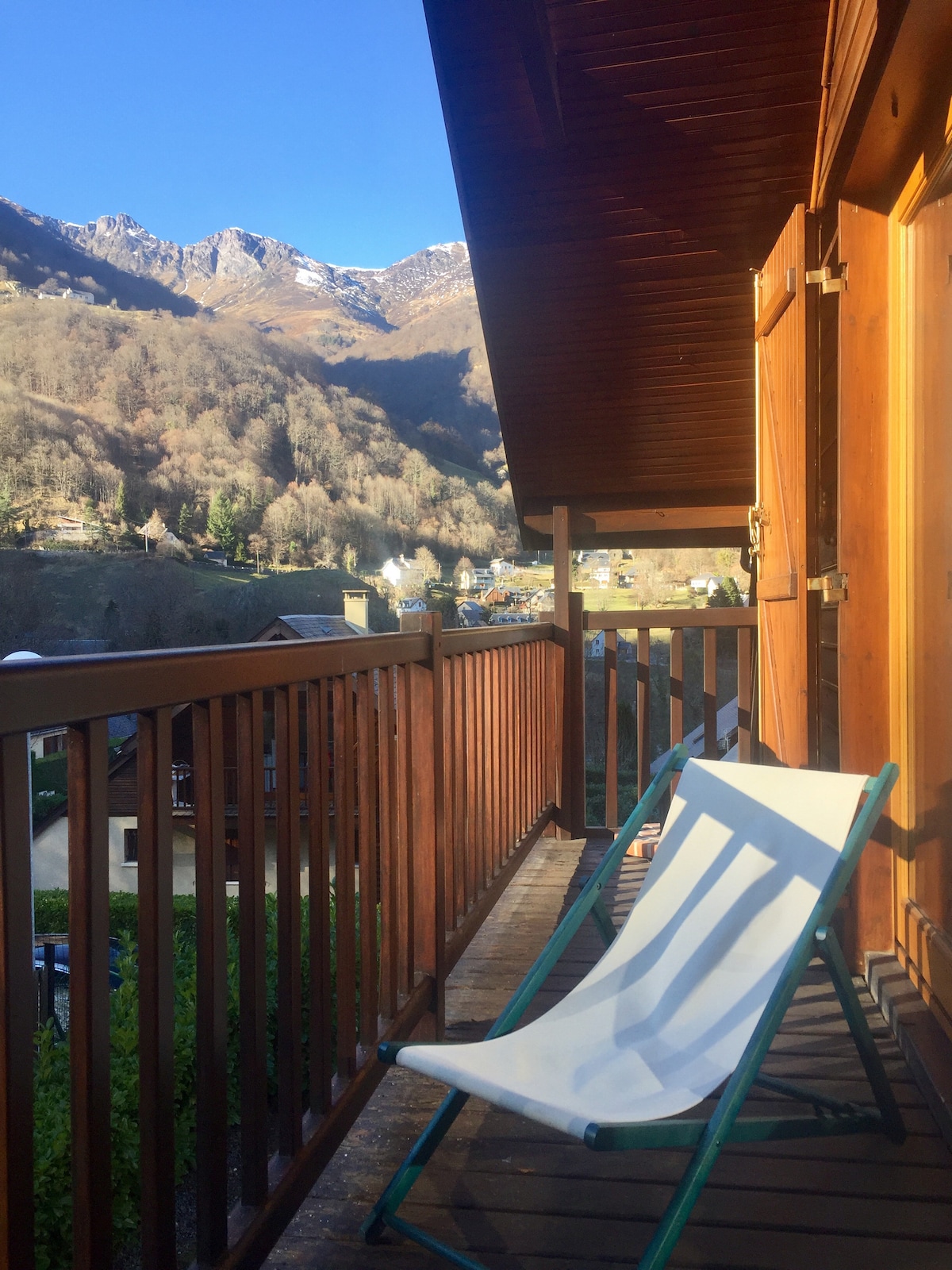 A wooden balcony extends outdoors, featuring a lounge chair positioned for relaxation. Scenic mountain views are framed by the surrounding trees, while bright sunlight illuminates the area, creating a serene outdoor space.