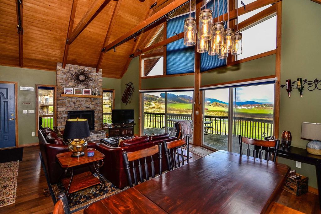 A spacious great room is shown with wooden beams and floor-to-ceiling windows. A stone fireplace is centered on the wall. Leather seating surrounds a wooden coffee table, while a dining table is visible nearby. Scenic mountain views are framed by large glass doors leading to the patio.