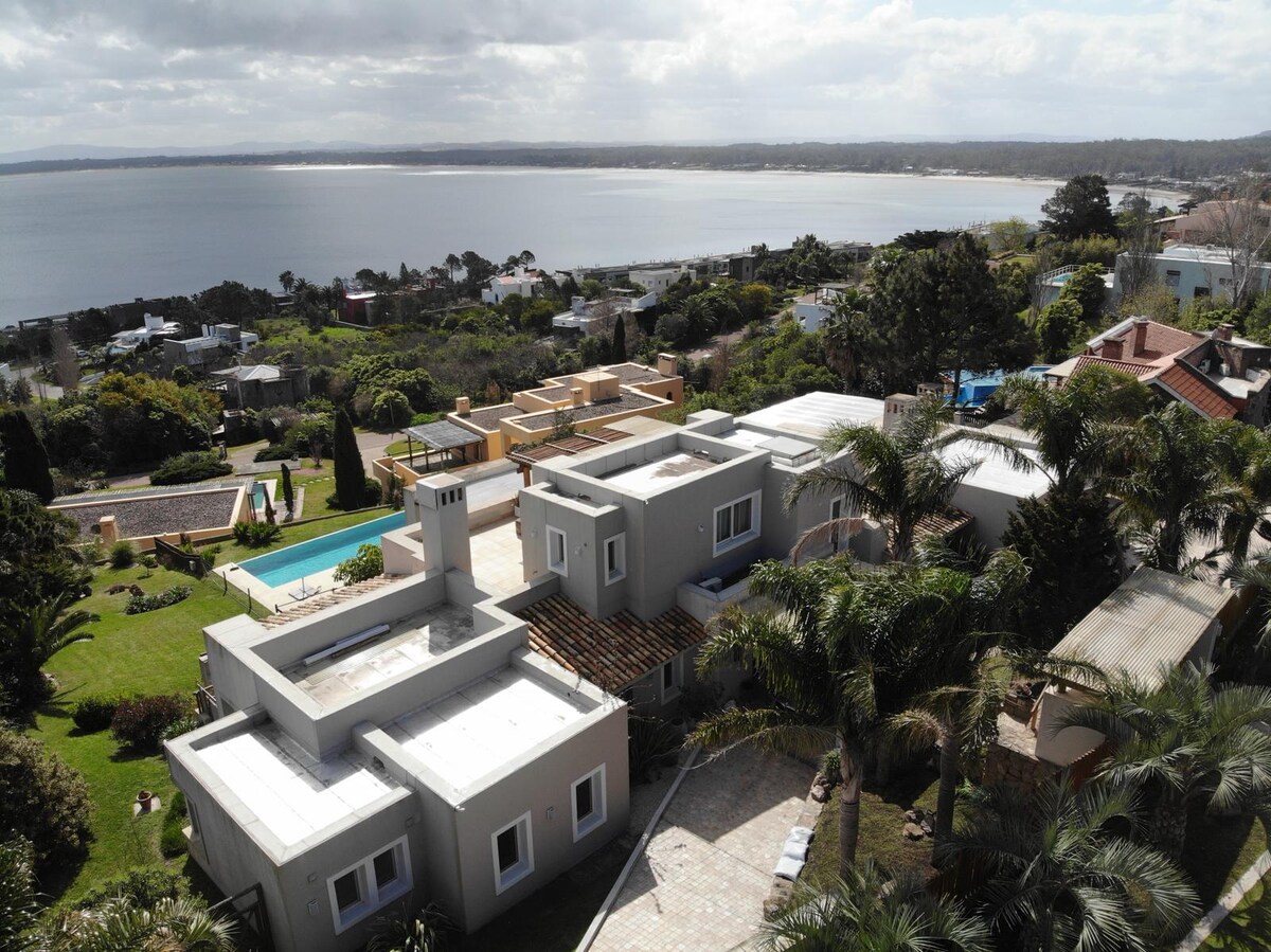An aerial view showcases a spacious guesthouse surrounded by lush greenery and a well-maintained garden. The house is positioned on a hillside, offering panoramic ocean views. Nearby, a shared pool glimmers under the afternoon sun, inviting relaxation amidst a tranquil landscape.