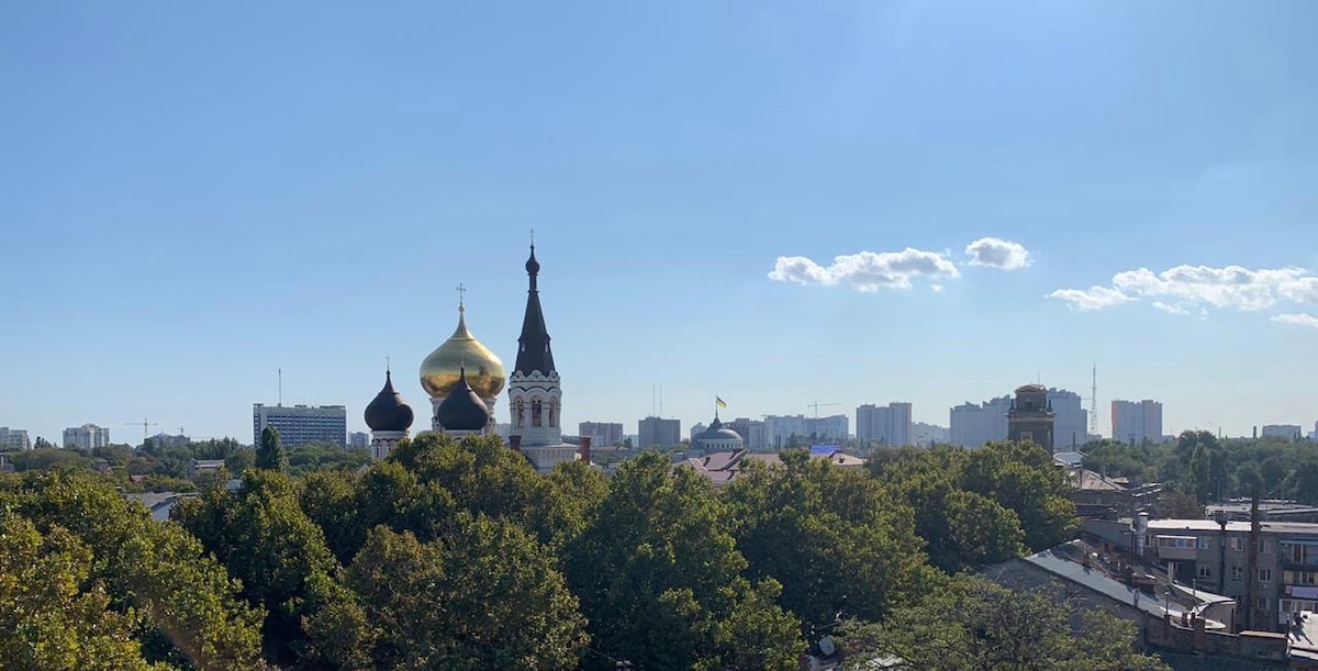A panoramic view showcases a skyline featuring distinct church domes, including a large golden dome. Lush green trees fill the foreground, contrasting with the city buildings in the background under a clear blue sky with scattered clouds.
