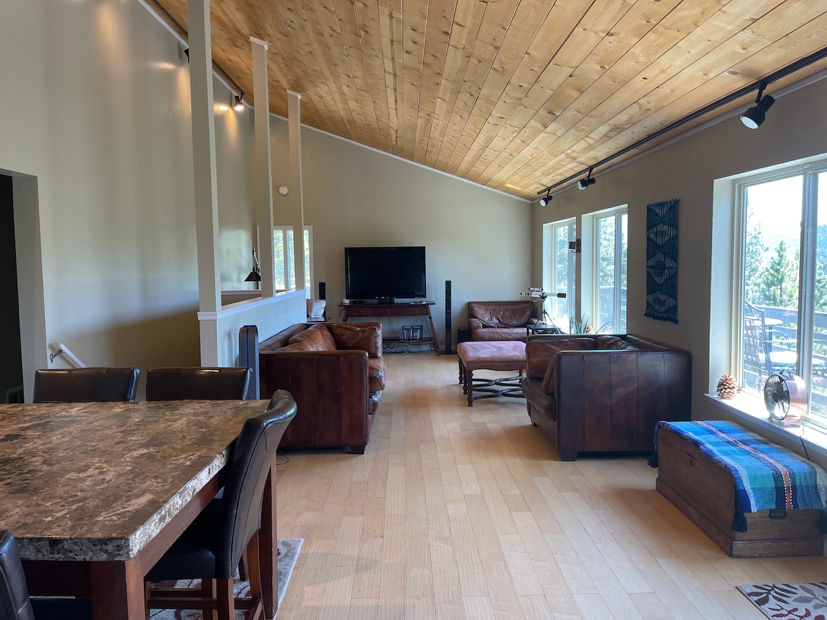 The spacious living and dining area features vaulted ceilings and large windows that provide natural light and views of the outdoors. Comfortable leather seating is arranged around a coffee table, with a dining table nearby. Light wooden floors enhance the open layout.