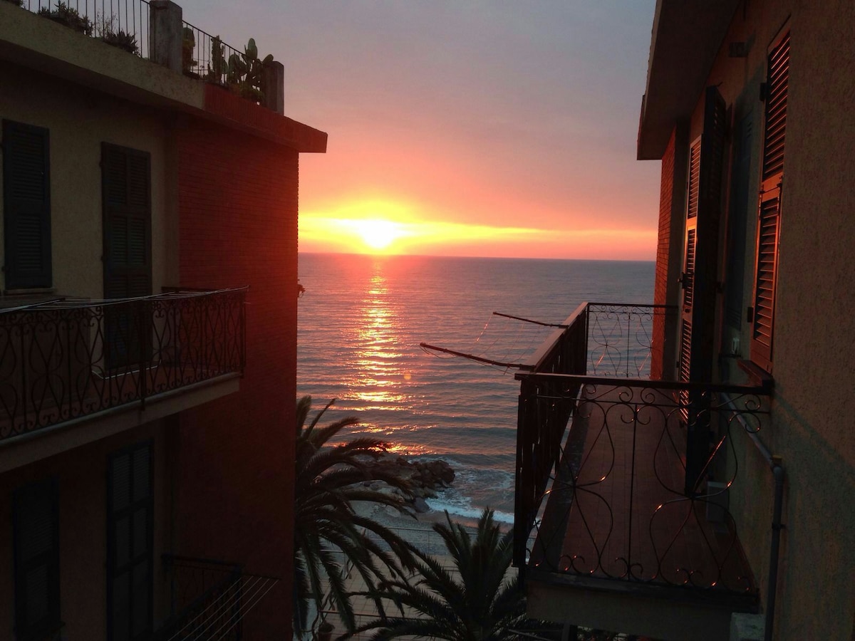 A sunset paints the sky in warm hues above the sea, seen from a balcony framed by nearby buildings. The sun reflects on the tranquil water, while palm trees line the shoreline, creating a serene coastal view.