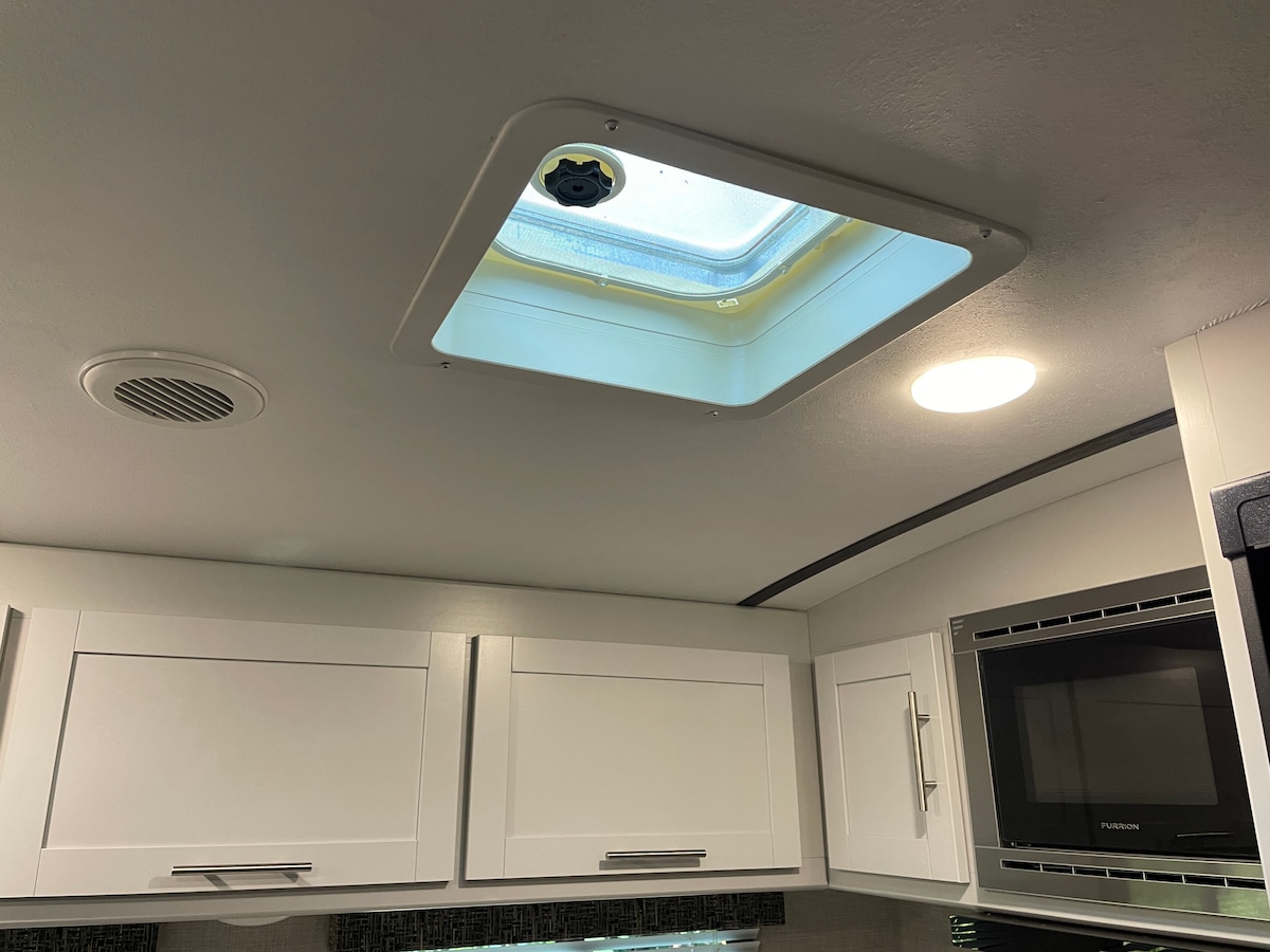 The interior of the camper is shown with a ceiling vent surrounded by white cabinetry. A circular light fixture is visible, providing illumination. The vent is partially open, allowing natural light to filter in.
