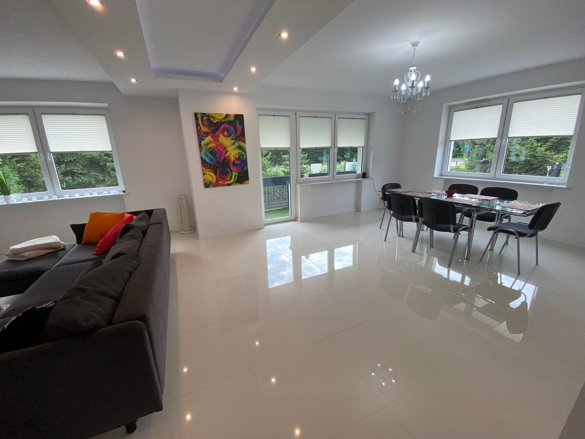 A modern living and dining area is showcased with glossy white flooring and ample natural light from large windows. A plush sofa in dark fabric complements a dining table set for six. An artistic wall piece adds a touch of color to the neutral decor.