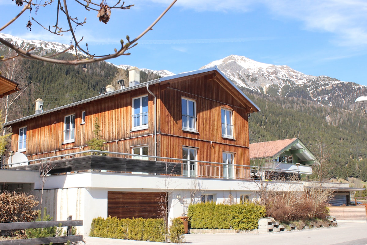 A wooden two-story house is presented with large windows, allowing natural light to illuminate the interior. A spacious balcony offers views of the surrounding mountains, while the exterior features landscaped greenery and a stone pathway leading to the entrance.