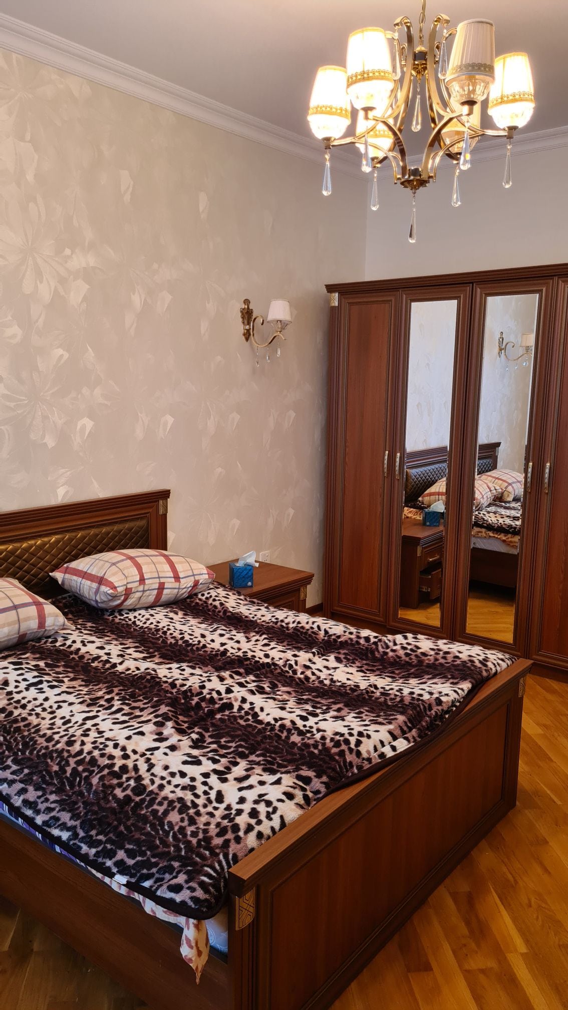 A cozy bedroom features a spacious bed adorned with a patterned blanket and decorative pillows. A wooden nightstand is positioned beside the bed, and a mirrored wardrobe reflects the soft lighting from a stylish chandelier overhead, contributing to a warm ambiance.