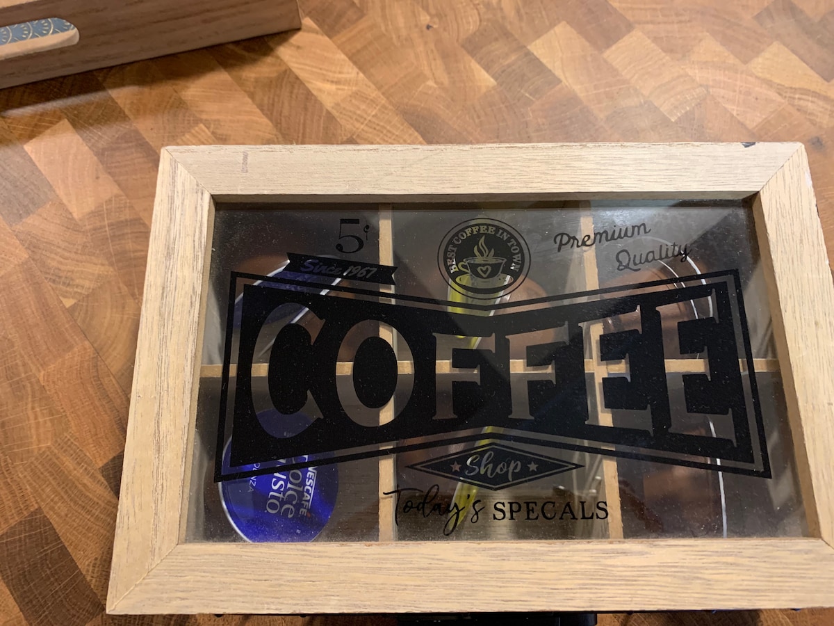 A wooden display case features a clear front showcasing various coffee capsules, each labeled with its flavor. The word 'COFFEE' is prominently displayed on the exterior, along with 'Today's Specials,' offering a welcoming touch for coffee enthusiasts.