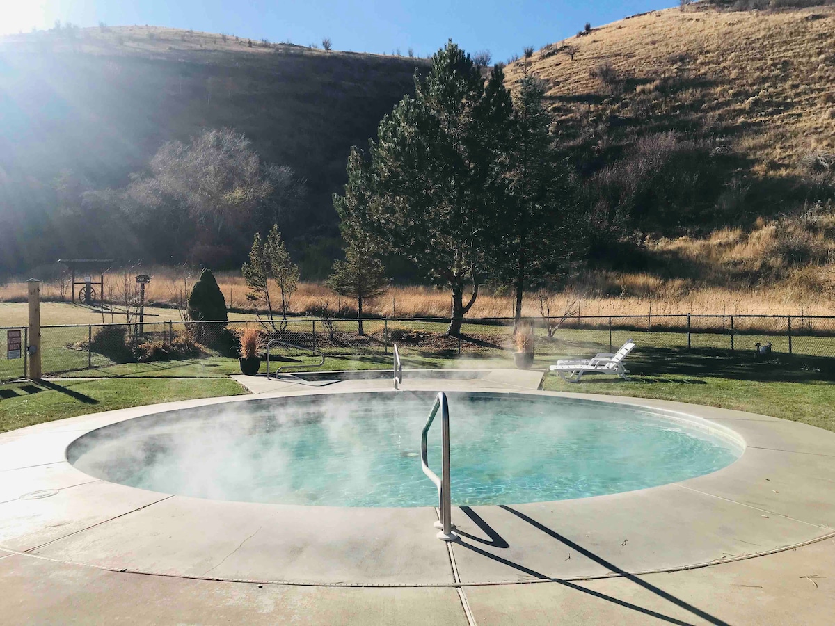 A circular hot springs soaking pool is shown with steam rising gently from the water, surrounded by a grassy area. Nearby, a few lounge chairs are positioned, and a fenced natural landscape features dry hills and sparse trees in the background under a clear blue sky.