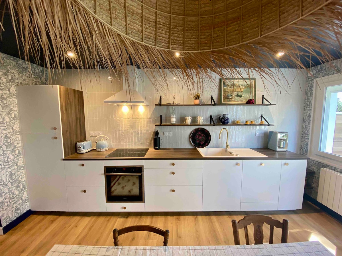A well-equipped kitchen features modern appliances, including an oven, sink, and coffee maker. Open shelving displays decorative items, while a woven ceiling creates an organic touch. The space is illuminated by natural light coming through a nearby window.