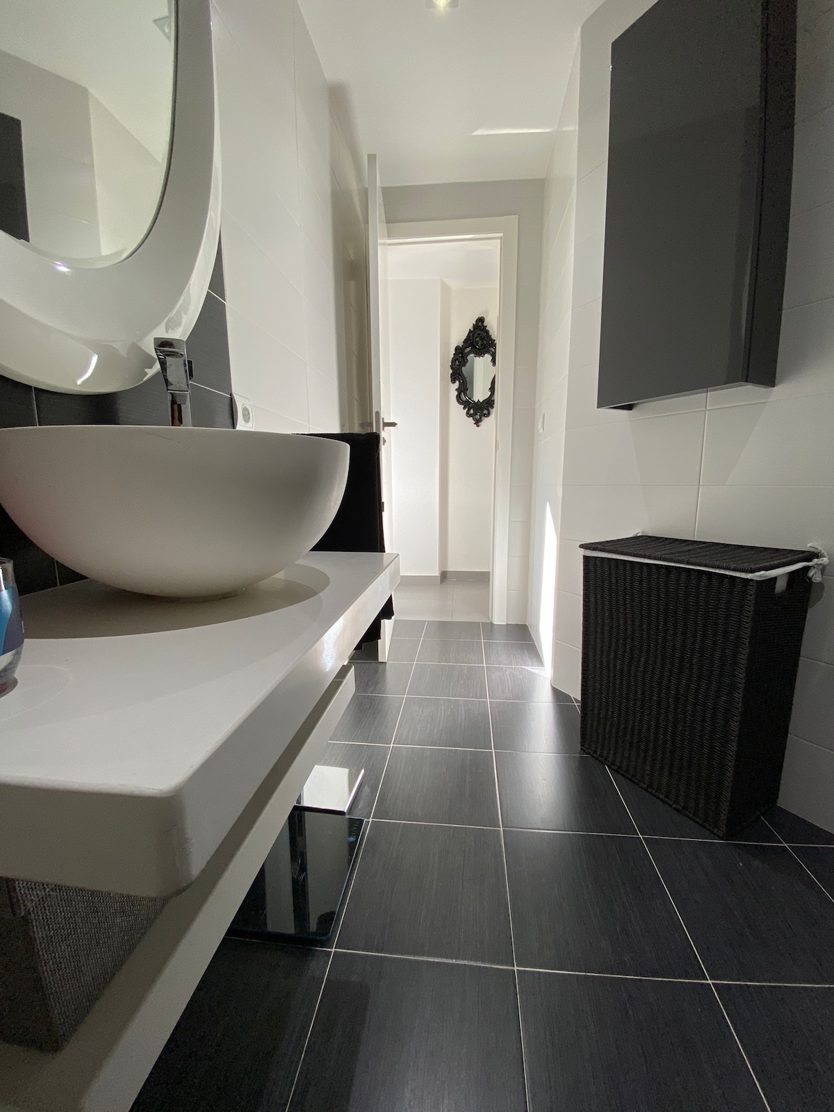 A modern bathroom is presented, featuring a sleek, round basin atop a white countertop. Dark tiles cover the floor, reflecting natural light. A mirror with an elegant frame adorns the wall, alongside a tall cabinet and a woven laundry basket, adding functionality to the space.