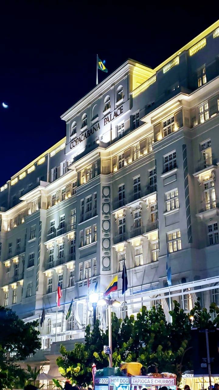 The illuminated facade of a historic building is displayed at night, showcasing a grand entrance framed by numerous columns and large windows. Flags from different nations are visible at the front, and the overall architecture reflects classic elegance with soft light highlighting the details.