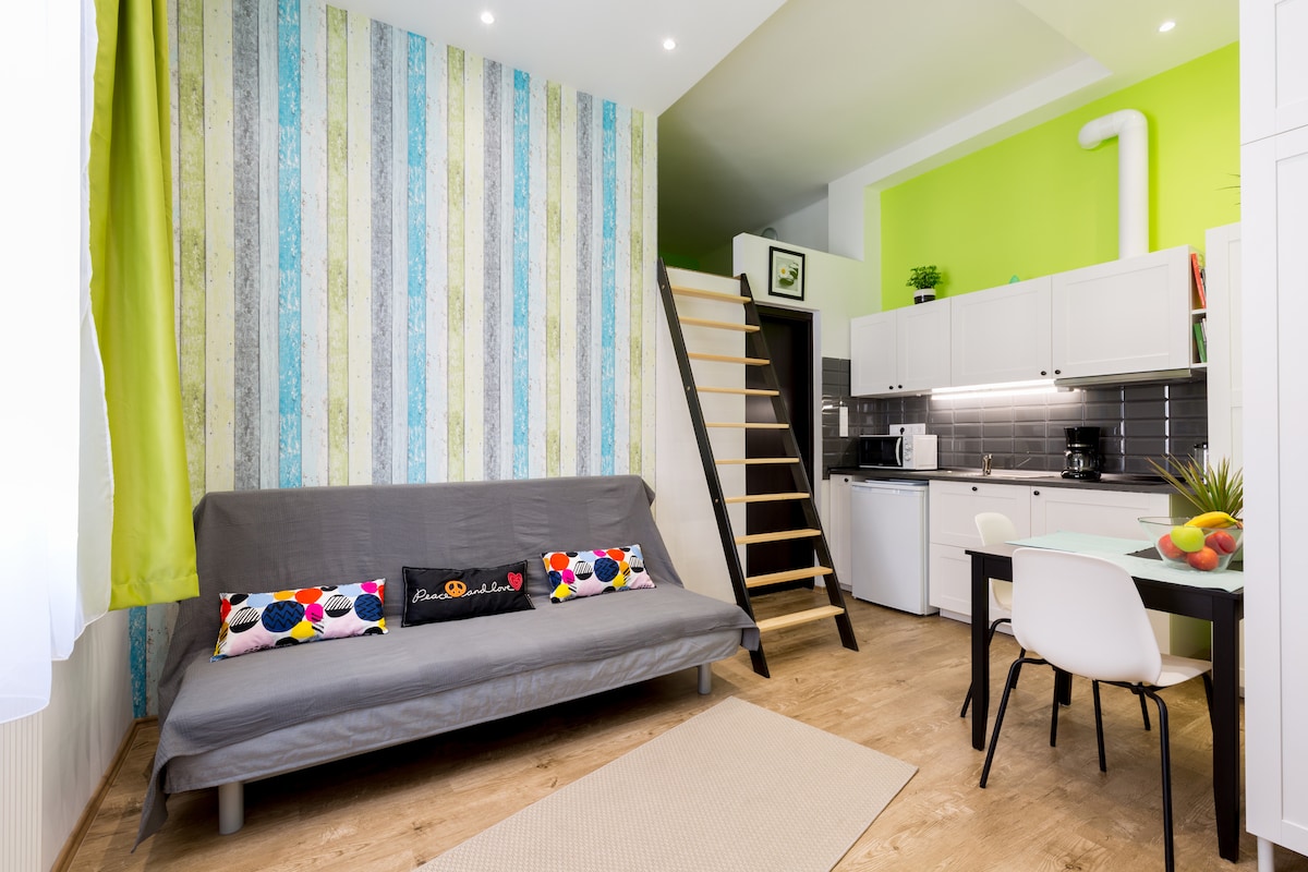 A modern studio features a colorful striped accent wall paired with a soothing green hue. A gray sofa bed provides flexible sleeping arrangements. A kitchenette is visible, equipped with essential appliances. A dining table is set for two, complemented by a small staircase leading to an upper sleeping area.