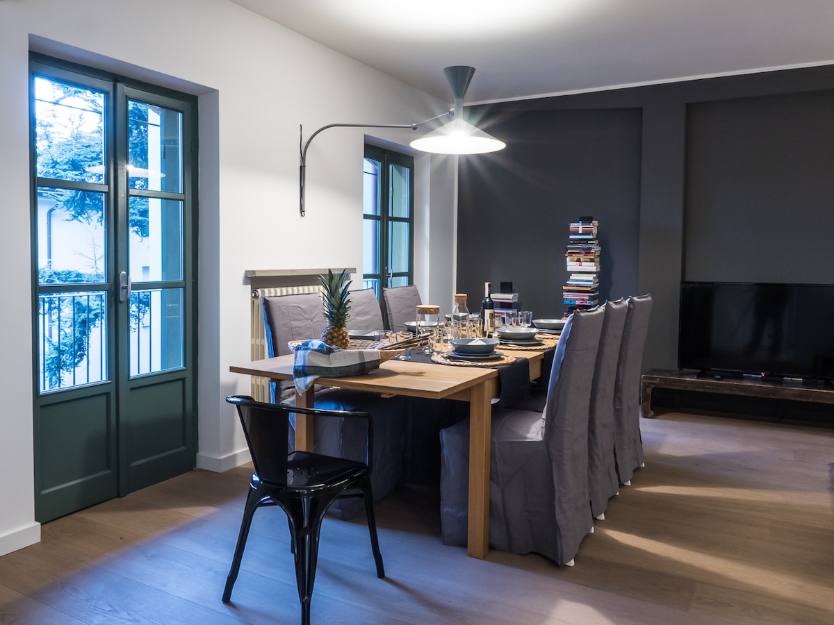 A dining area features a large wooden table set for a meal, surrounded by upholstered chairs. Soft light is provided by a modern pendant lamp. Two large windows allow natural light, with green shutters framing the view, while a television and bookshelf add to the space.