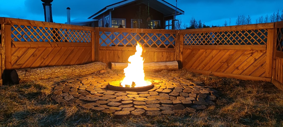 A circular fire pit made of stone is surrounded by a wooden enclosure with lattice work. Flickering flames dance at the center, providing warmth in the evening. The cabin's silhouette is visible in the background against a twilight sky.