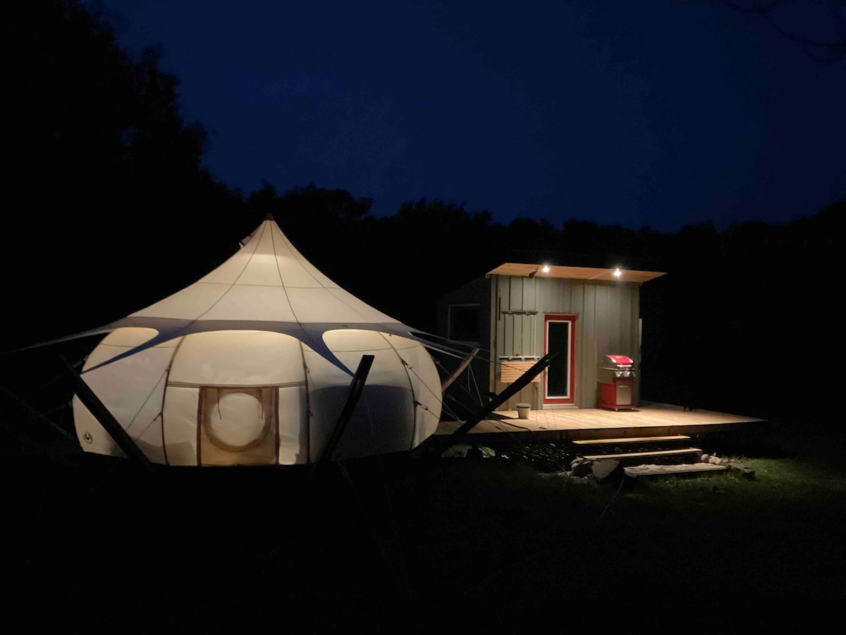 A spacious Lotus Belle tent is illuminated at night, highlighting its unique shape. Adjacent, a private kitchen with a red grill and a wooden deck can be seen. Soft shadows create a serene atmosphere in the surrounding darkened landscape.
