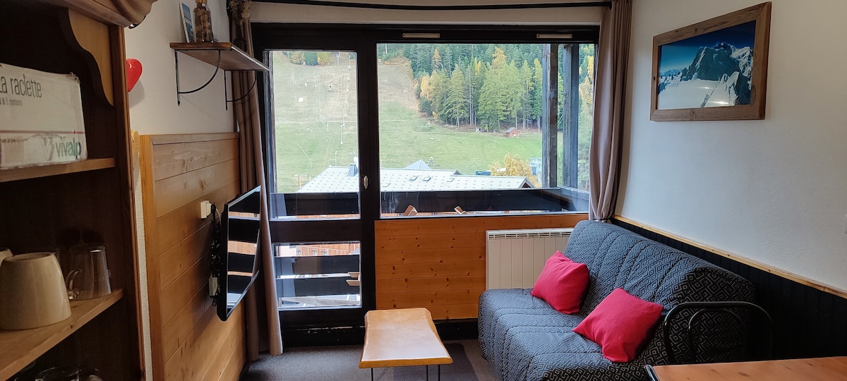 A cozy living area is presented with a gray sofa adorned with red cushions. A wooden coffee table is positioned in front of a large bay window, offering views of the surrounding greenery and slopes. A television is mounted on the wall, complementing wooden accents.
