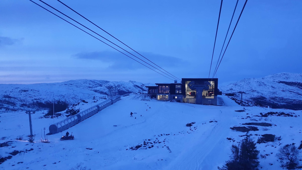 A modern building is set against a snowy landscape, illuminated by soft evening light. Skiers can be seen traversing the sloped terrain, while ski lifts ascend to the nearby slopes. The surrounding mountains create a serene backdrop.