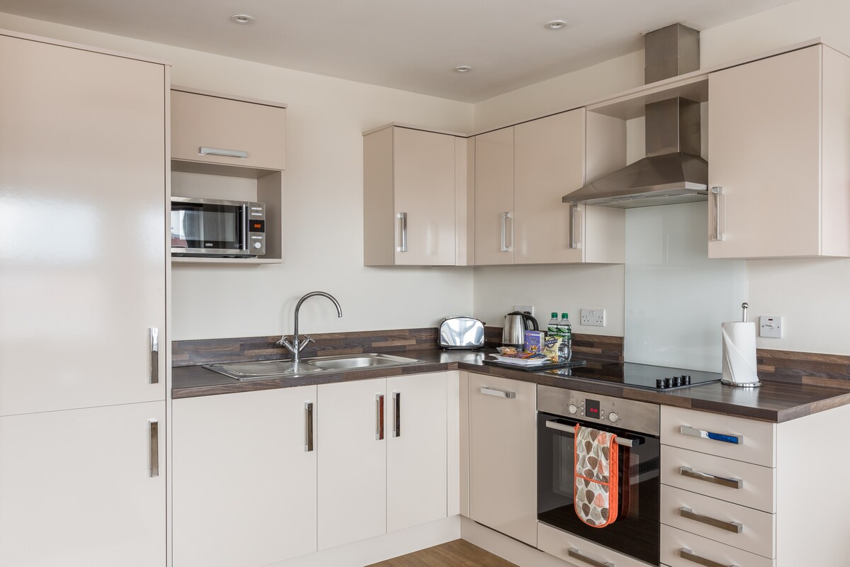 A modern kitchen area is depicted, featuring light-colored cabinetry and a dark countertop. Appliances include a microwave, kettle, and an oven with a gas hob. A small selection of kitchenware is neatly arranged, contributing to an organized and functional space.