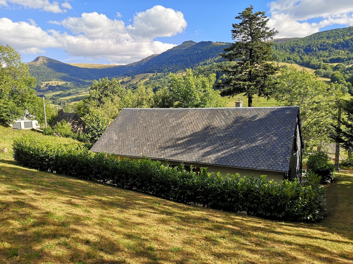 A modern building is set within a grassy area, surrounded by lush greenery. Scenic mountains rise in the background under a partly cloudy sky, creating a natural setting. The landscape is open and inviting, showcasing the tranquil environment of the location.